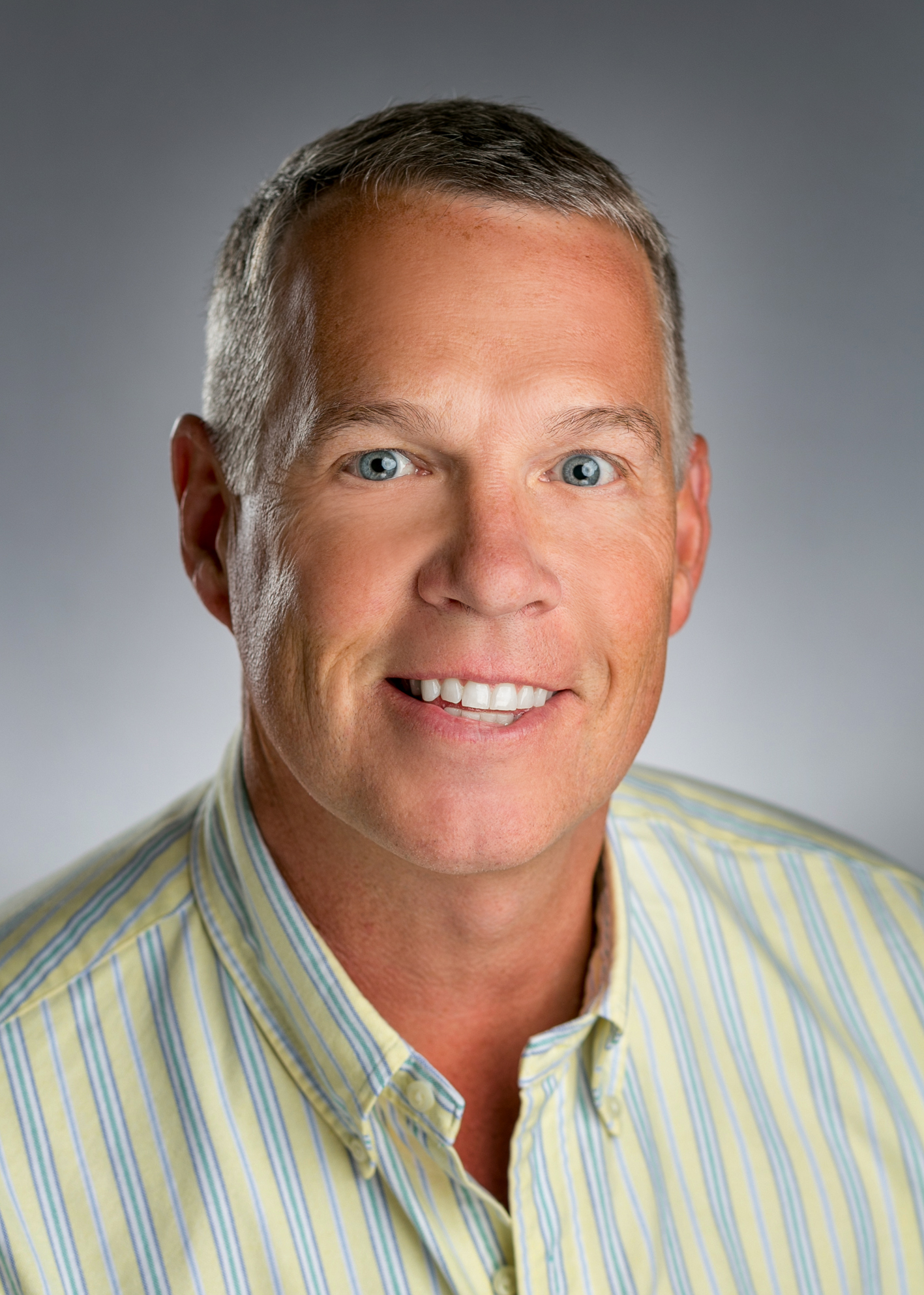 The image shows a man with short hair smiling at the camera against a neutral background.