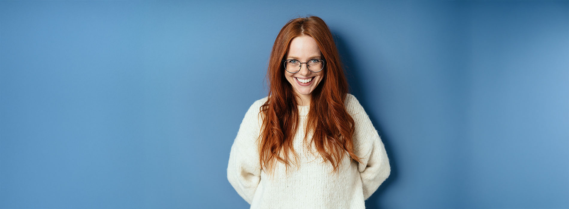 The image shows a person with long red hair standing against a blue background, wearing a white sweater and glasses, looking directly at the camera with a neutral expression.