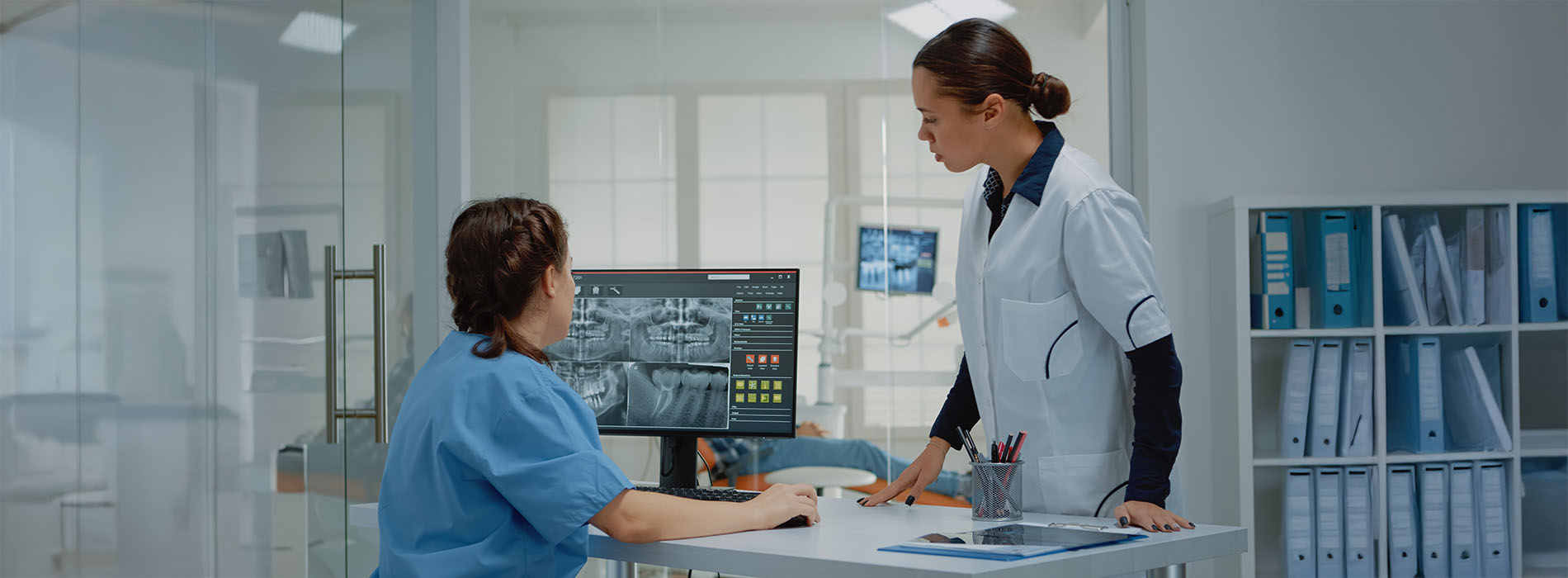 A woman in a white coat stands at a desk with a computer monitor, while another person, possibly a patient or visitor, looks on from behind.