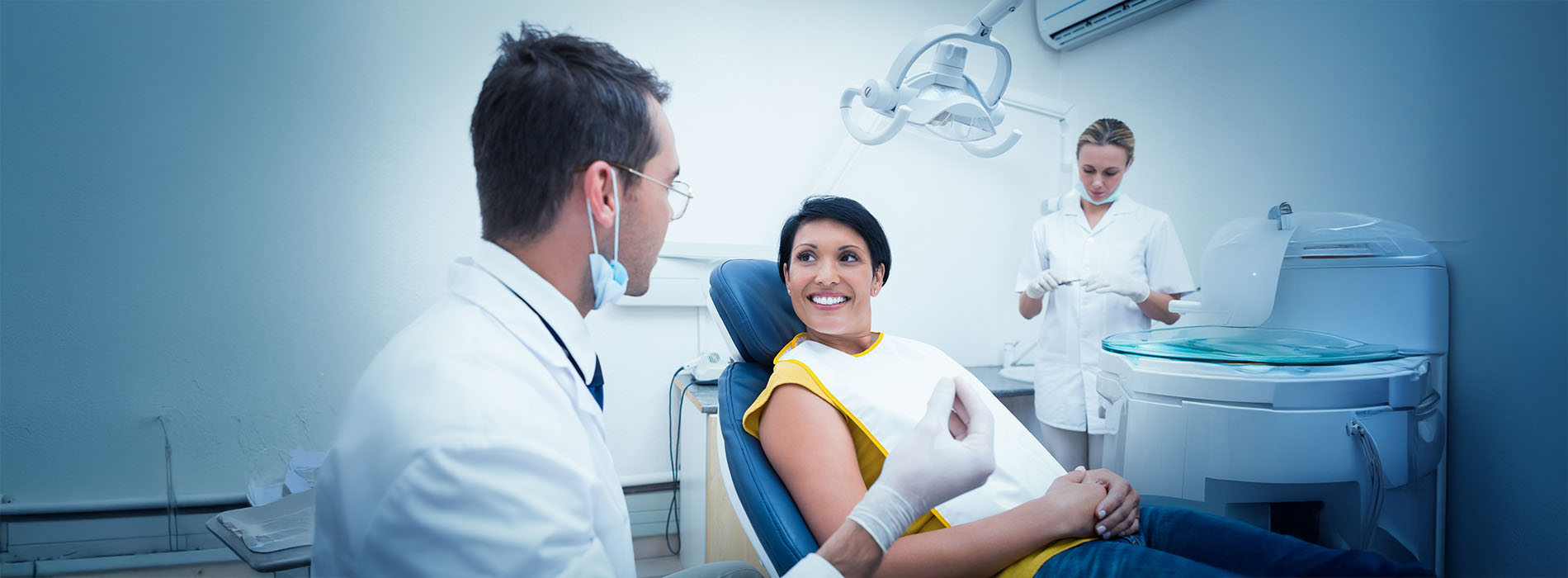 The image depicts a professional dental office setting with a dentist and patient, where the dentist is seated at a chair and the patient is seated on a dental chair, both wearing protective face masks, and the scene suggests an ongoing dental examination or treatment.