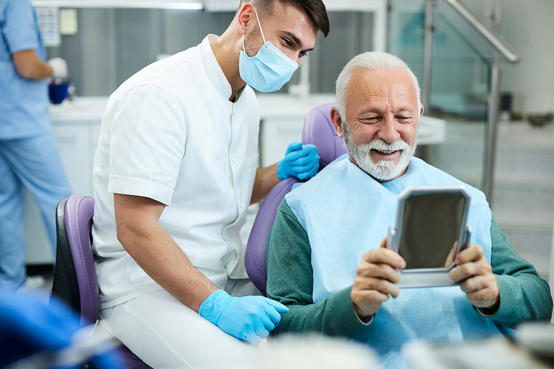 In the image, there s a dentist wearing a white lab coat and a surgical mask, sitting at a dental chair with a patient who has a beard, holding a tablet. The setting appears to be a modern dental clinic.