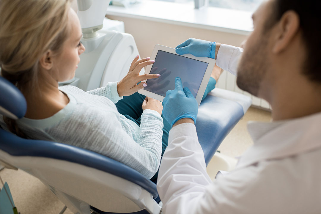 A dental professional demonstrates a digital device to a patient in a dental office chair.