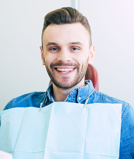 The image shows a smiling man with a beard sitting in a dental chair wearing a blue shirt and a white mask, looking directly at the camera.