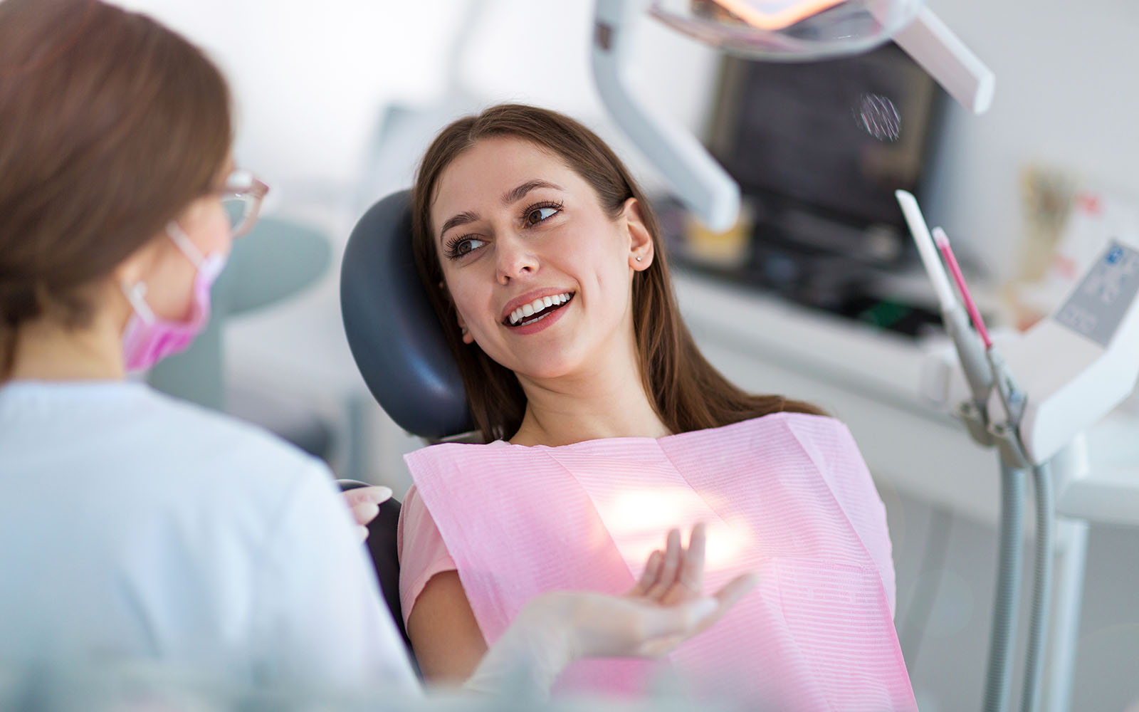 A woman sitting in a dental chair with a smile on her face while a dentist looks at her teeth.