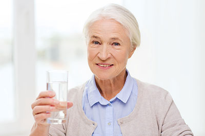 An elderly woman holding a glass of water with both hands while smiling at the camera.