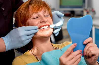 A woman wearing a red mask is seated in a dental chair with her mouth open, receiving dental care from a professional who is adjusting her teeth using tools.