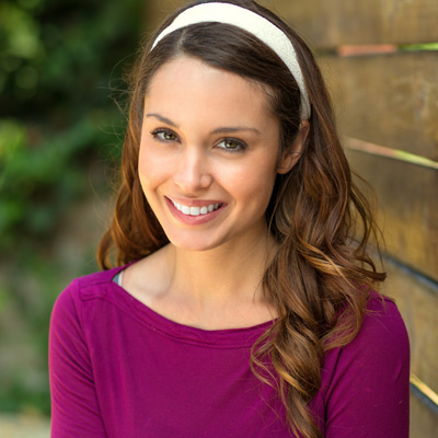 A smiling woman with long hair, wearing a purple top, stands against a wooden fence.