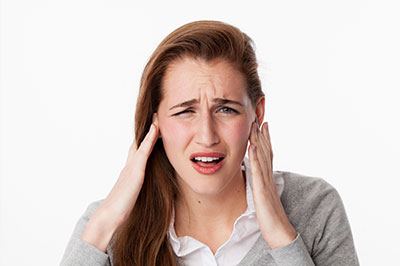 The image shows a woman with her hand on her head, appearing to be experiencing discomfort or pain, possibly related to her ears, against a white background.