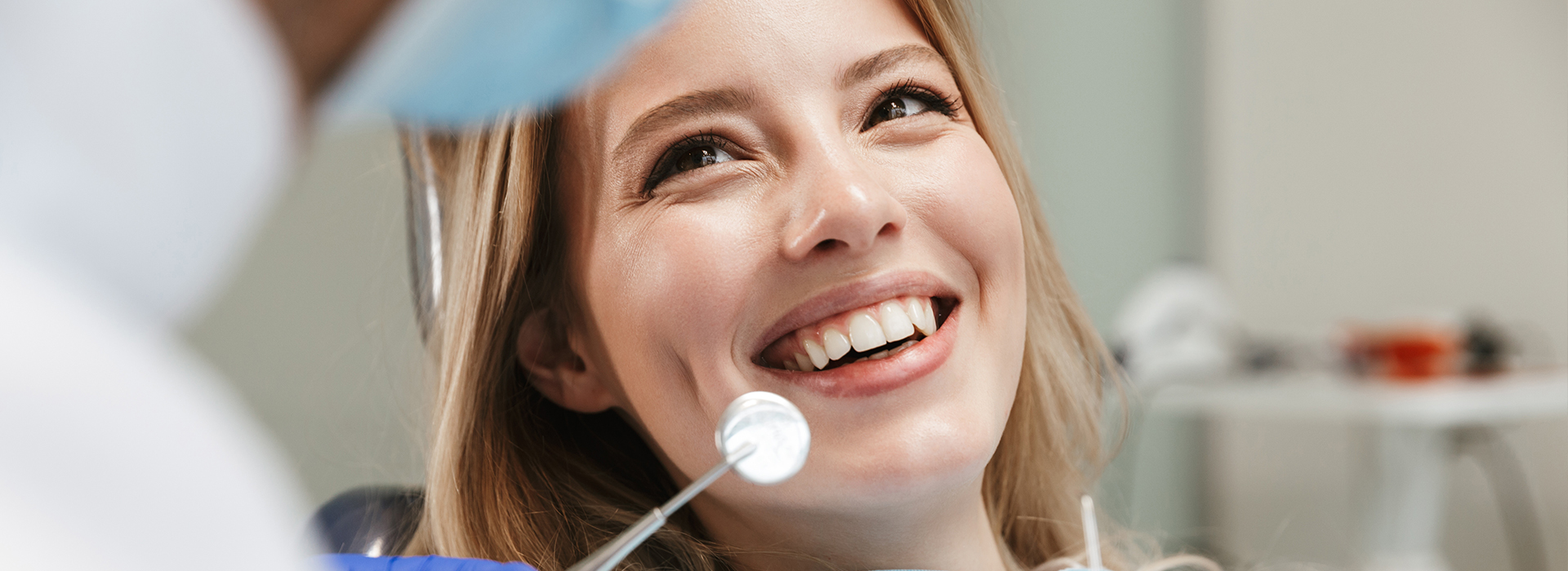 A woman smiling at a camera while seated in a dental chair with a dentist in the background.