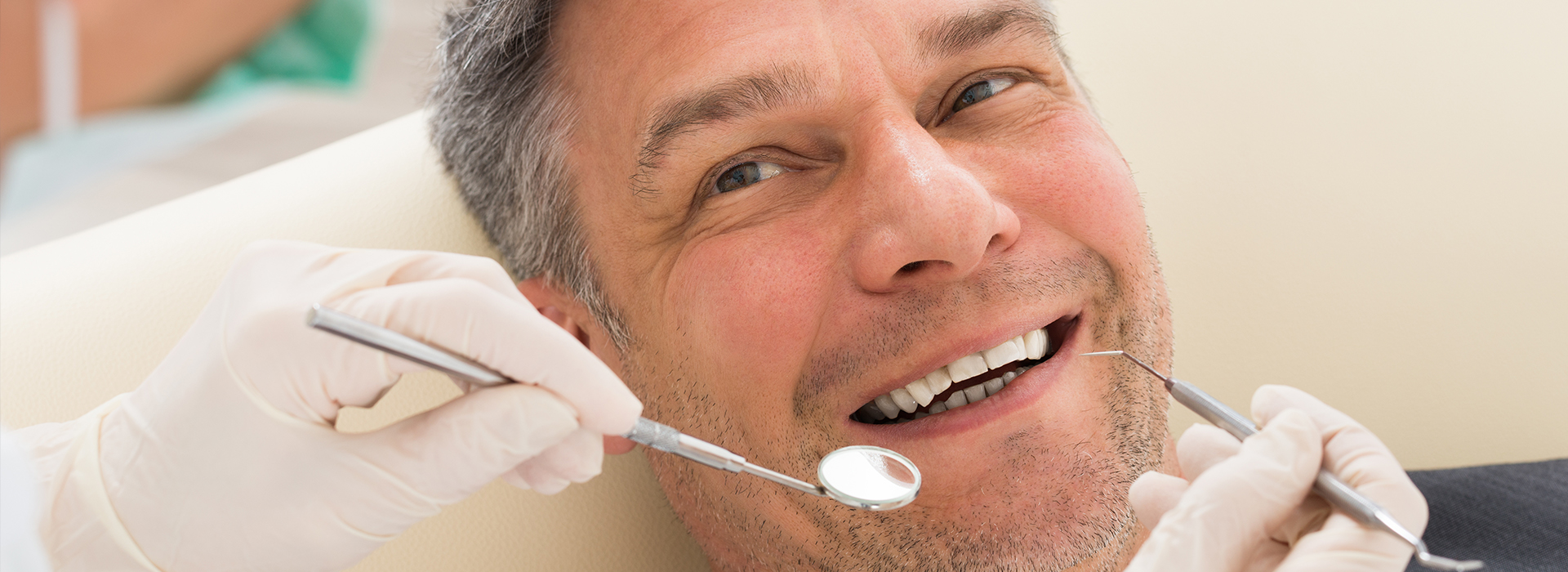 The image shows a man with a toothy smile sitting in a dental chair while receiving dental care, with a dentist working on his teeth using dental instruments.