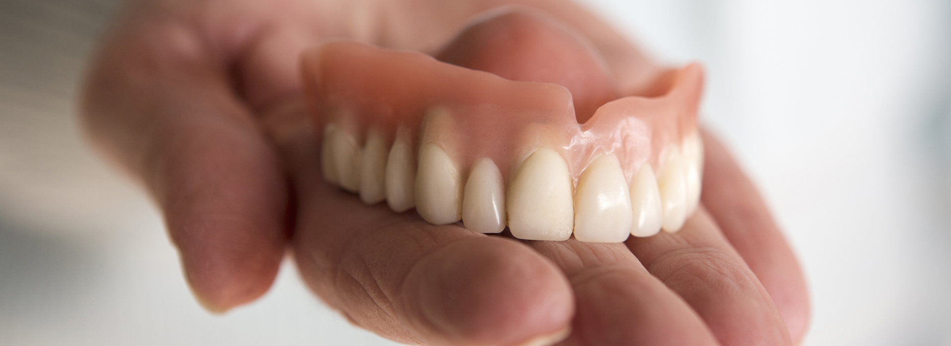 The image shows a hand holding an artificial set of dentures against a blurred background.