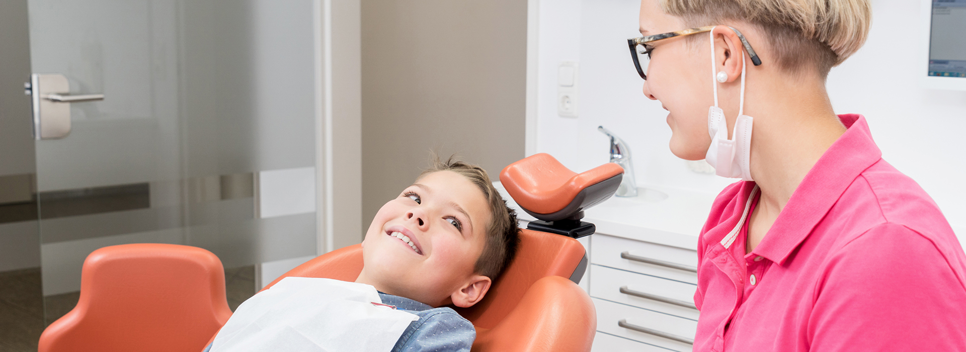 A photograph depicts a dental office setting with an adult seated in a chair having their teeth checked by a dentist, who is standing beside them holding a device close to the patient s mouth.