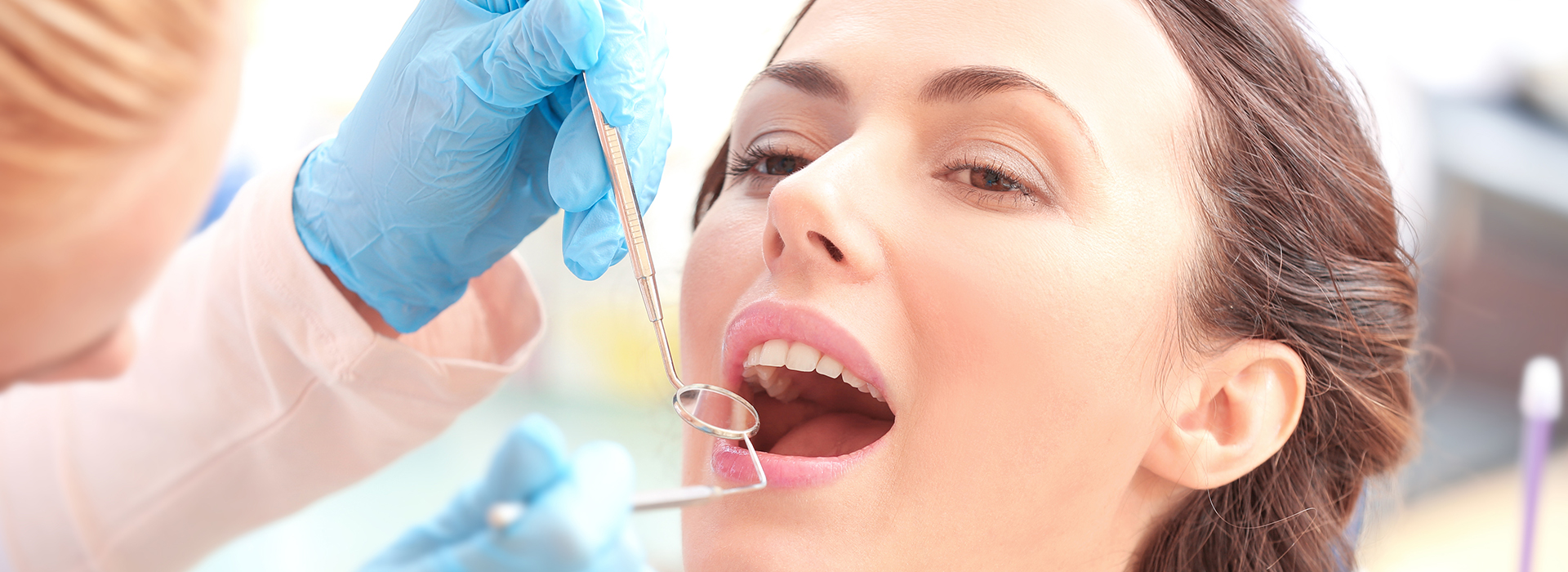 A woman receiving dental treatment with a dental hygienist performing the procedure, both wearing protective gloves and masks.