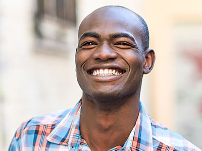 A smiling man with short hair, wearing a plaid shirt, stands outdoors against a backdrop of a building with a window and a red door.