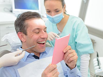 A man sitting in a dental chair, holding a pink card with a smile on his face, while a dentist looks at him with a surgical mask and tools around them.