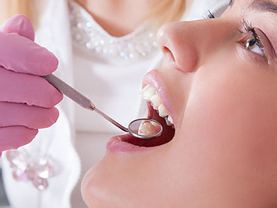 A dental hygienist is performing a cleaning procedure on a woman s teeth using an ultrasonic scaler, while she wears protective eyewear and a pink gown.
