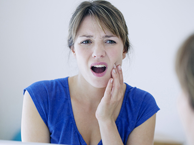 A woman with her mouth open wide, looking at her reflection in a mirror, appears surprised or concerned.