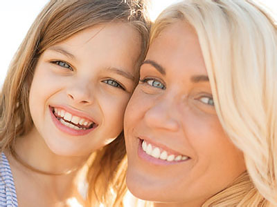 A photograph of a woman with blonde hair and a young girl standing beside her, both smiling at the camera.