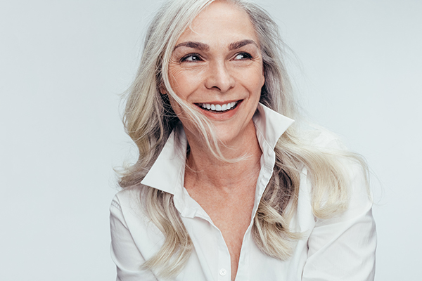 The image shows a woman with blonde hair smiling at the camera. She has her hair pulled back and is wearing a white top. Her expression conveys warmth and approachability.