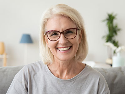 The image shows a woman with short blonde hair, wearing glasses and a light-colored top, smiling at the camera while seated indoors.