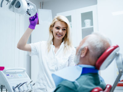 A woman in a white coat stands next to an elderly man seated in a dental chair, both in a dental office setting.