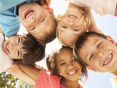A group of children smiling at the camera, with their arms around each other, set against a clear sky background.