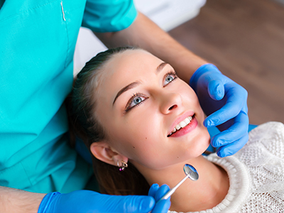 A dental hygienist is performing a cleaning procedure on a woman's teeth while she smiles at the camera, both are wearing gloves and protective eyewear.