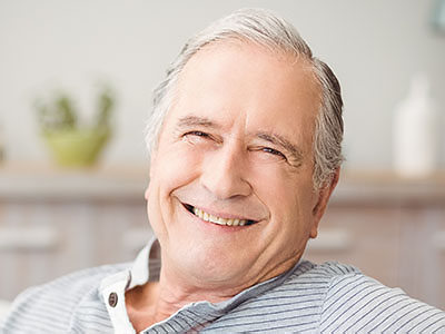 The image shows an elderly man with white hair and glasses, smiling at the camera while sitting in a living room.