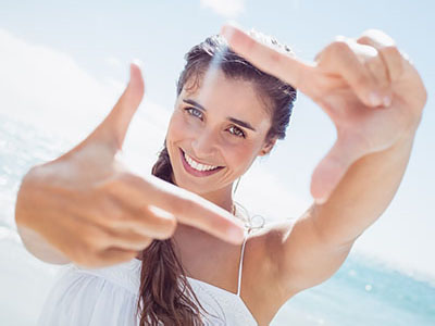 A woman with long hair is smiling while taking a selfie, set against a beach backdrop.